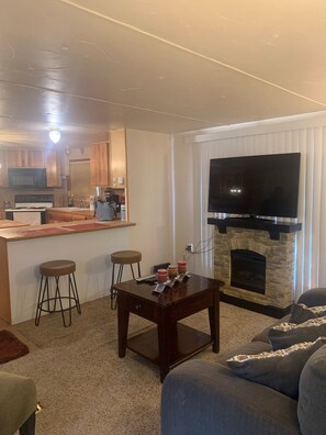 A living room with a fireplace, a TV, and a kitchen area with wooden cabinets and a bar.