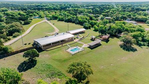 An aerial view of a property with a large building, a swimming pool, and surrounding green fields. - Cedar Creek Ranch on Lake Whitney (Whitney)