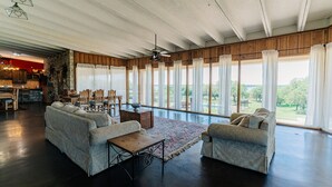 A spacious living room with a large window, a ceiling fan, and a kitchen area visible in the background. - Cedar Creek Ranch on Lake Whitney (Whitney)