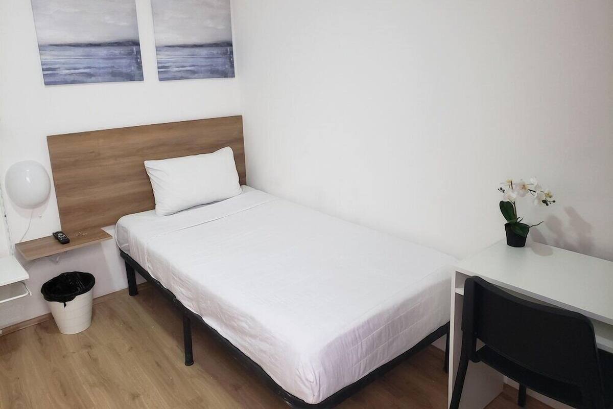 Un lit simple avec une tête de lit en bois, une table de chevet avec une lampe, une petite poubelle, un bureau avec une chaise et deux tableaux accrochés au mur.