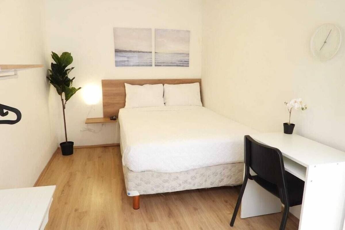 Une chambre à coucher comprenant un lit, une table de chevet, une lampe, une plante en pot, un bureau, une chaise et une horloge murale.