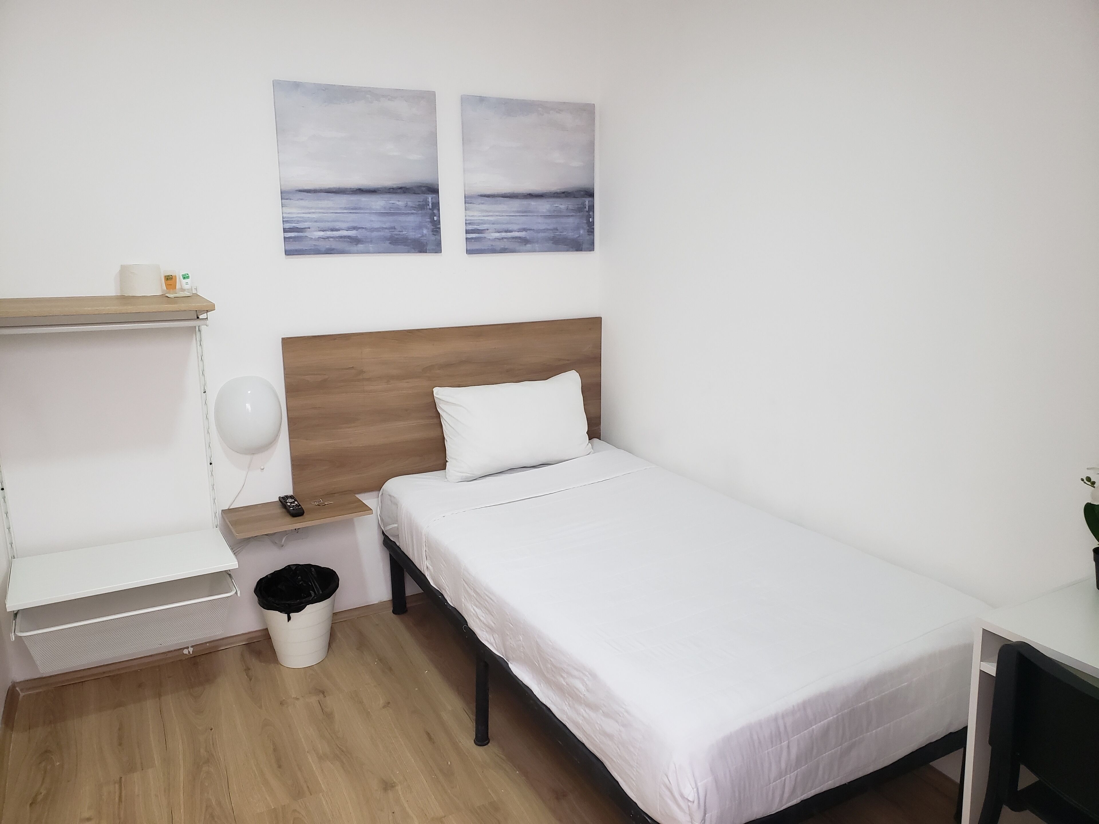 A minimalist bedroom with a wooden headboard, white bedding, a small bedside table with a lamp, and two abstract paintings on the wall.