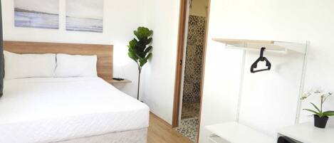 Un dormitorio con cama, escritorio, silla, perchero y planta en maceta.