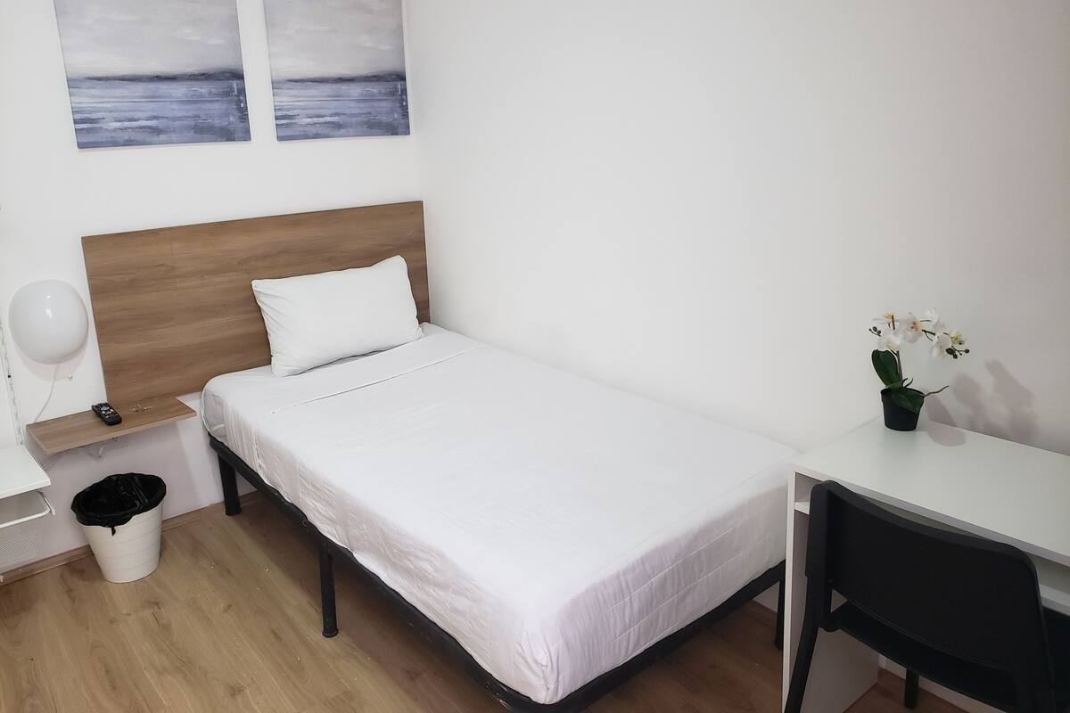 Une chambre à coucher bien rangée, avec une tête de lit en bois, un lit simple avec une literie blanche, une petite table de chevet avec une lampe, une poubelle, un bureau avec une chaise et deux tableaux accrochés au mur.