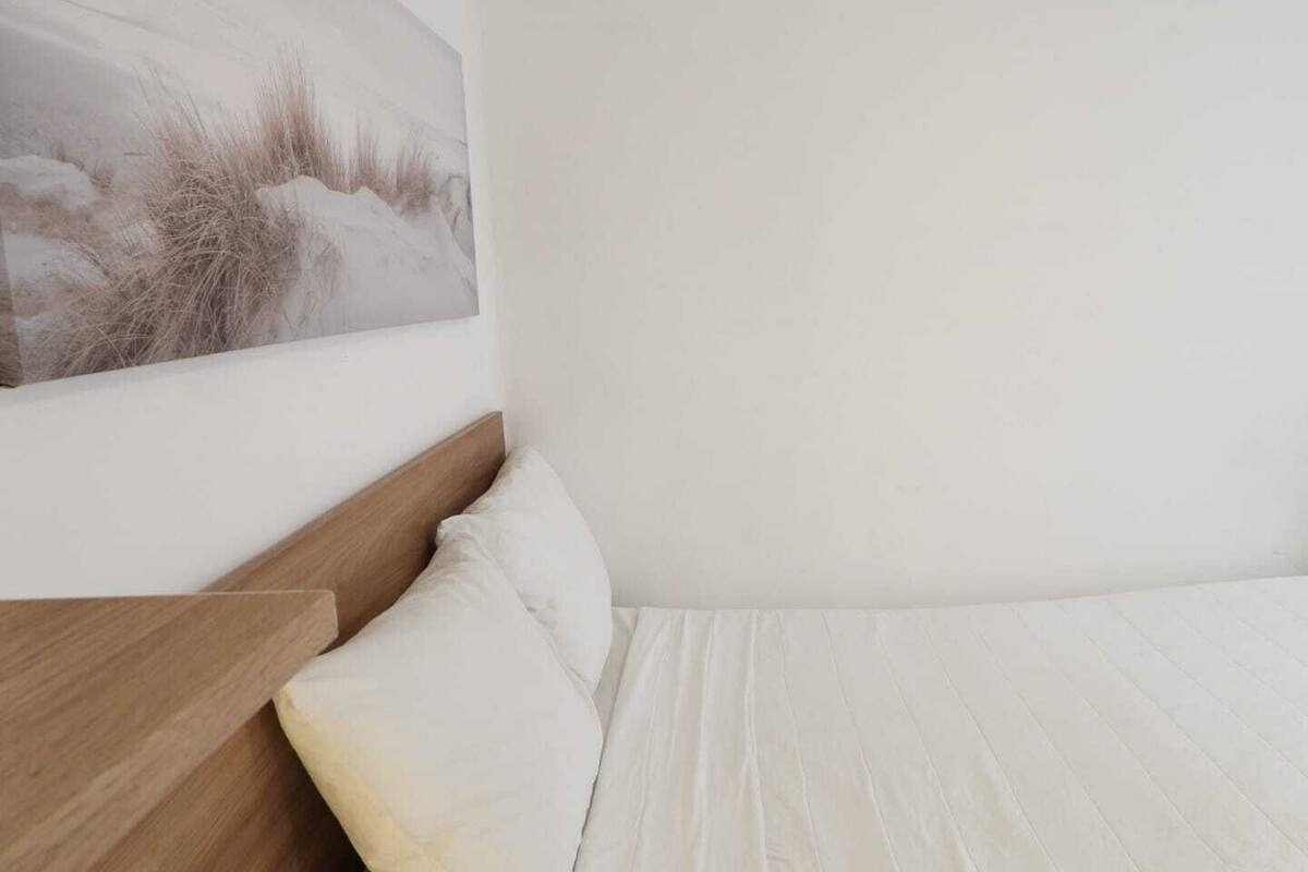 Une chambre à coucher avec une tête de lit en bois, une couvre-lit blanche et un tableau représentant un paysage enneigé accroché au mur.