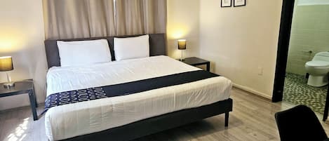 Habitación de hotel ordenada con una cama grande, mesitas de noche, una silla y vistas al baño.