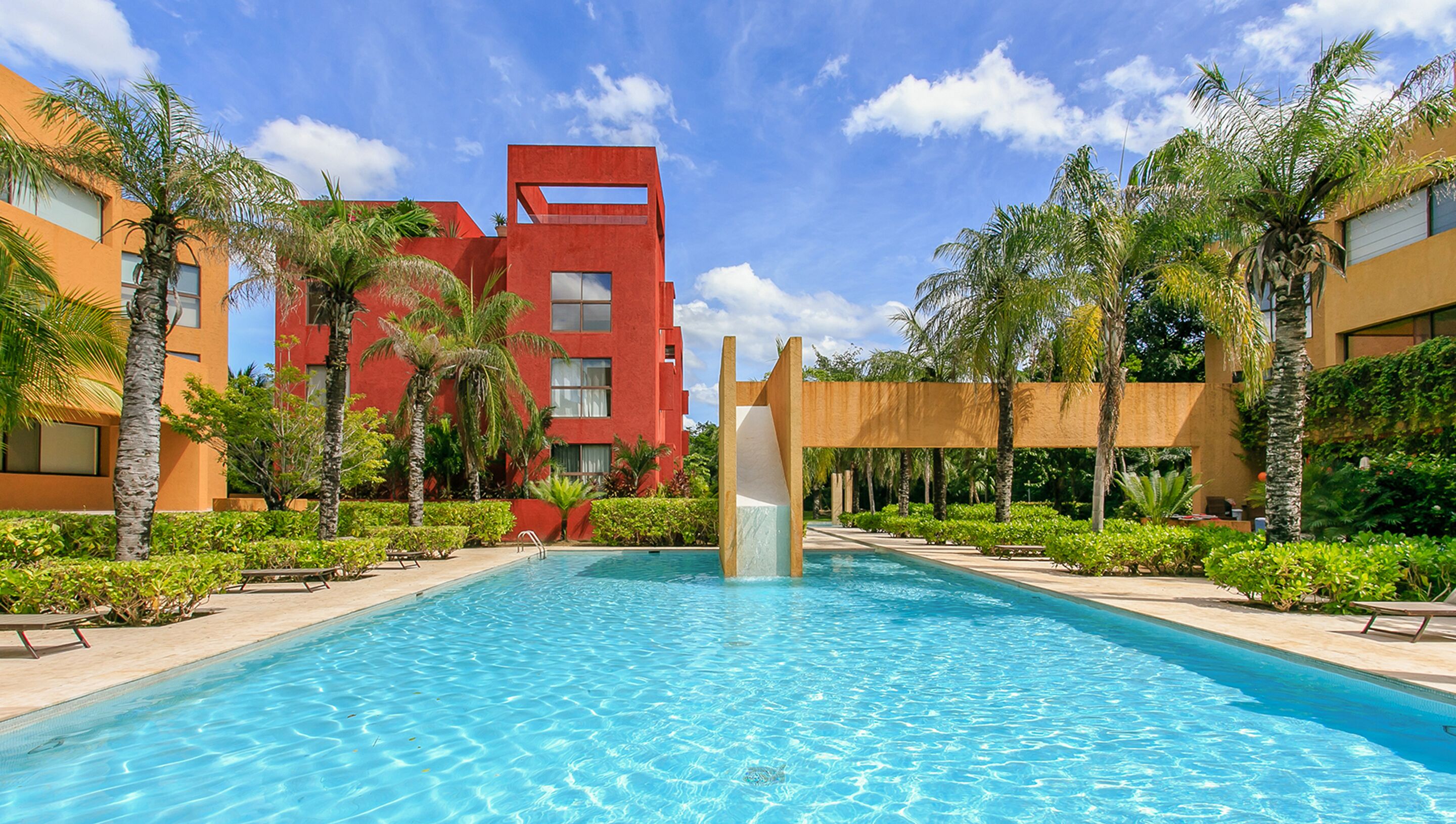 Una piscina con tobogán, rodeada de palmeras y un edificio rojo.