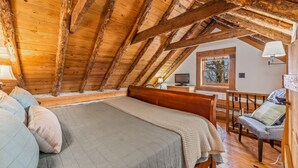 Un dormitorio acogedor en el ático con techo de madera, cama con almohadas, escritorio con lámpara y ventana con vistas al exterior.
