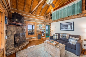 L’intérieur chaleureux d’un chalet avec une cheminée en pierre, des poutres en bois, un canapé confortable et un téléviseur.