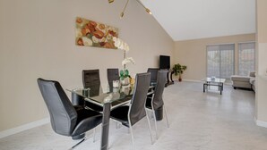 A dining area with a glass table, black chairs, a gold chandelier, and a floral painting on the wall.