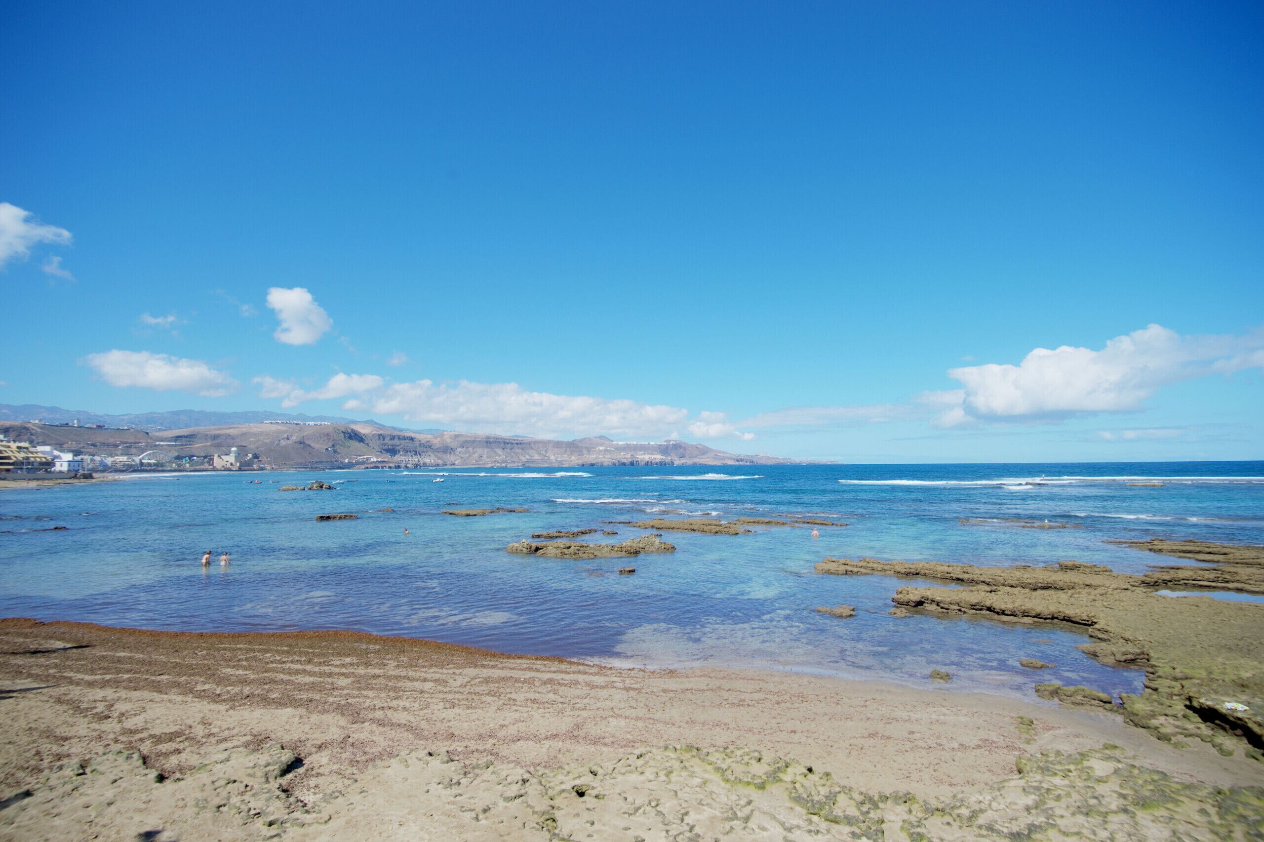 A coastal beach with clear blue water, rocky formations, and distant hills.