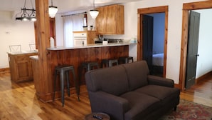 A modern kitchen with wooden cabinets, a bar area with stools, and a living room with a sofa and a remote control on a barrel.