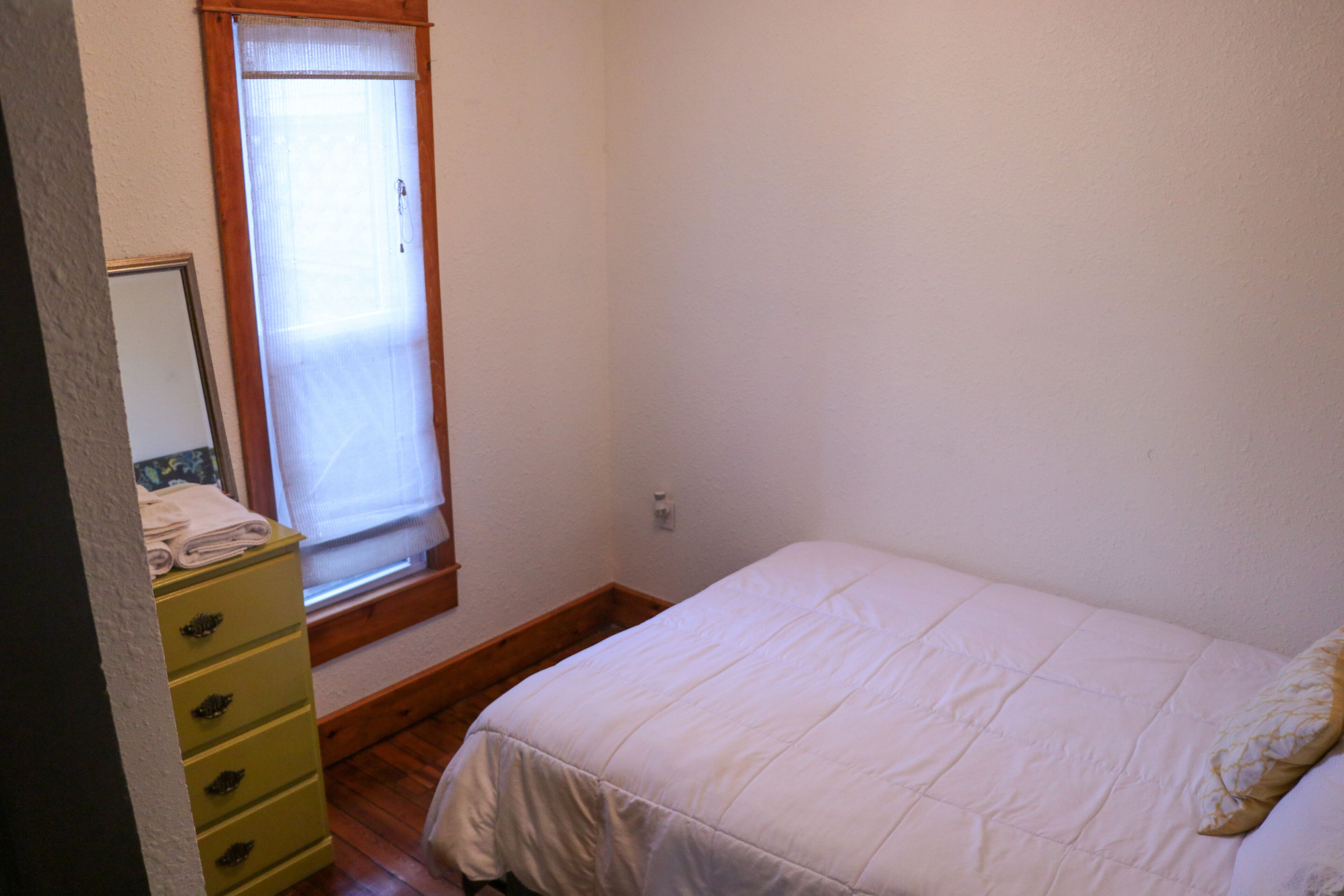A bedroom with a bed, a window with a curtain, a wooden dresser, and a wall-mounted mirror.