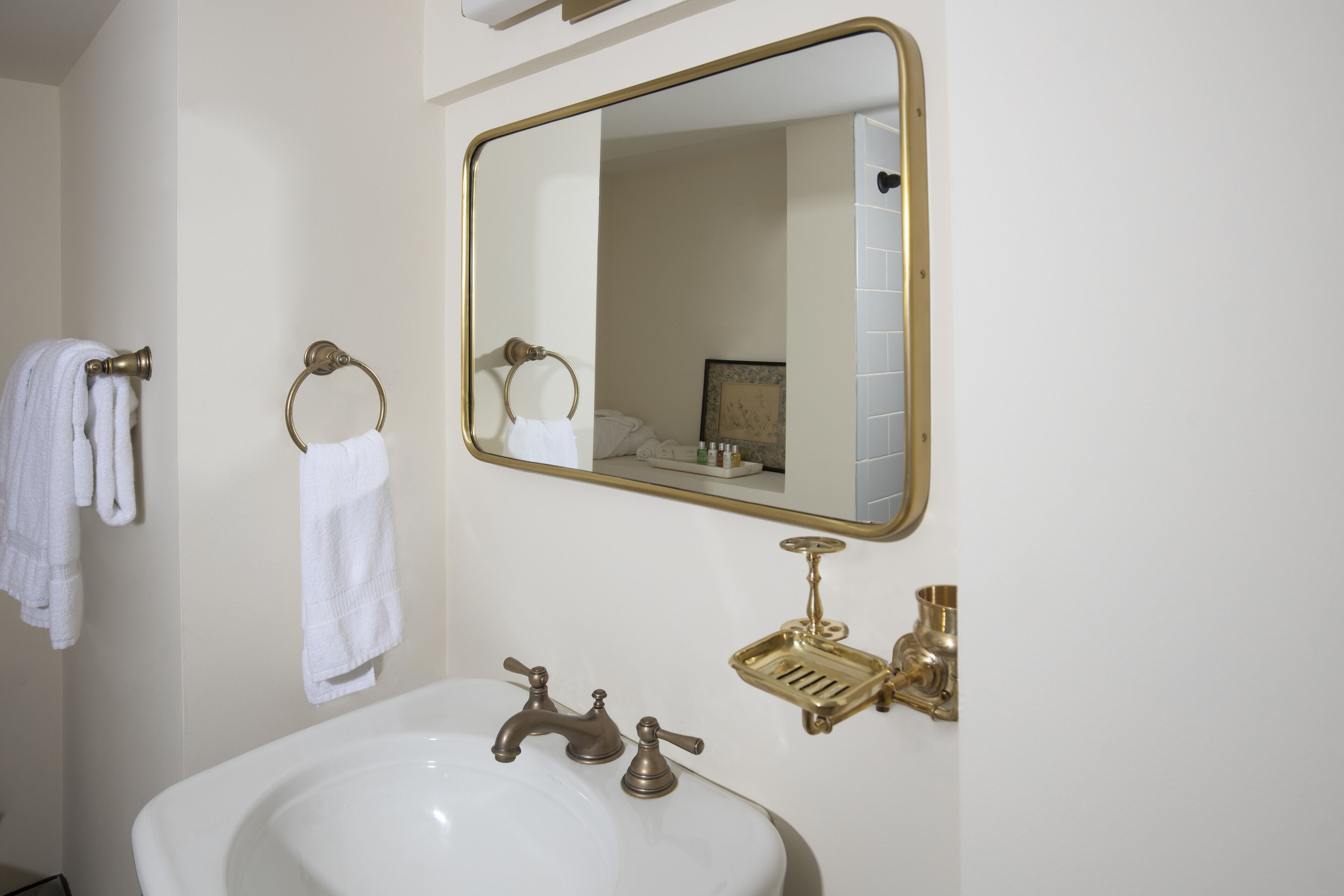Une salle de bain avec un lavabo blanc, des robinetteries en laiton, un miroir à cadre doré et des serviettes suspendues à un porte-serviettes.