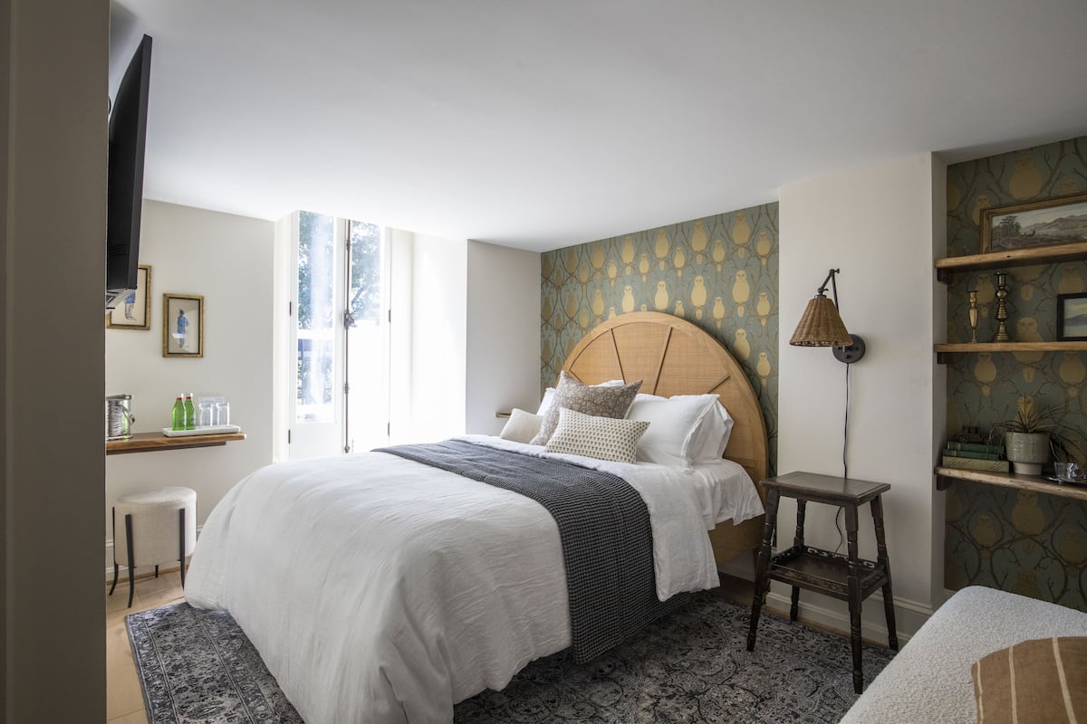 A neatly arranged bedroom with a large bed, a bedside table, a lamp, and a shelf with decorative items.