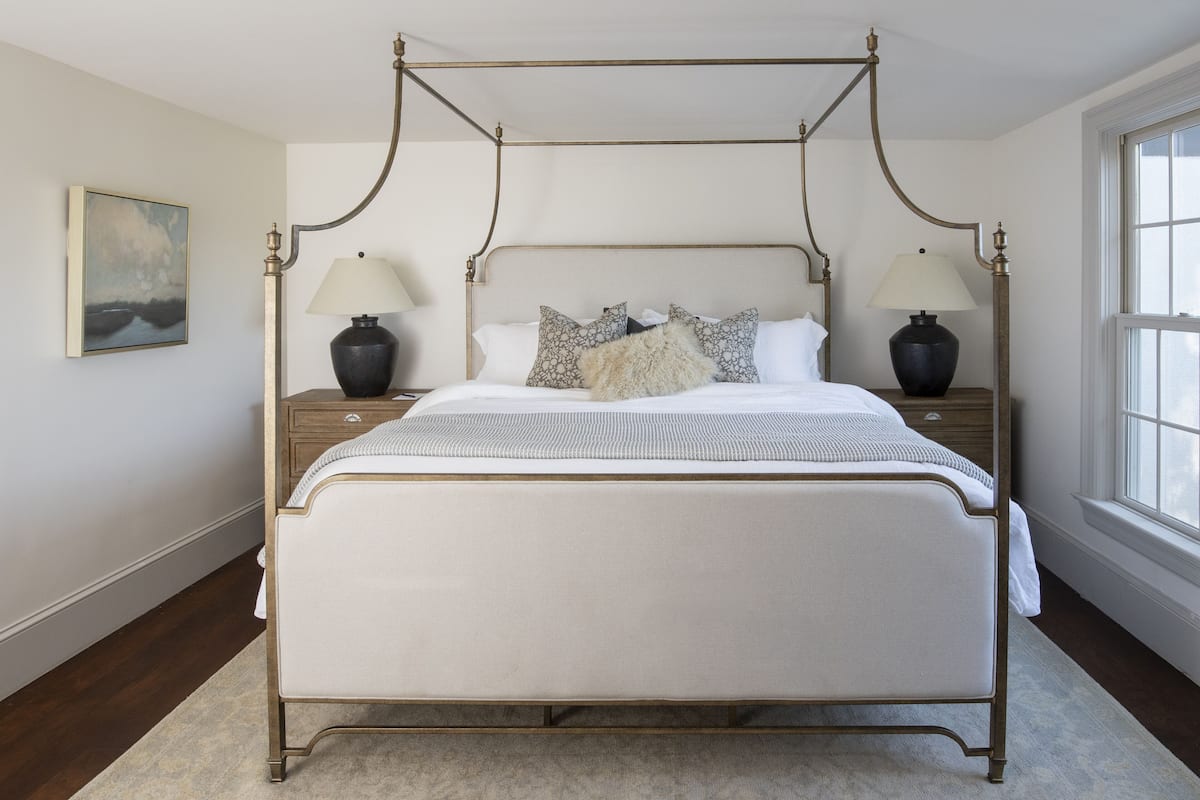 A four-poster canopy bed with a white mattress, two bedside tables with lamps, and a large window on the right.