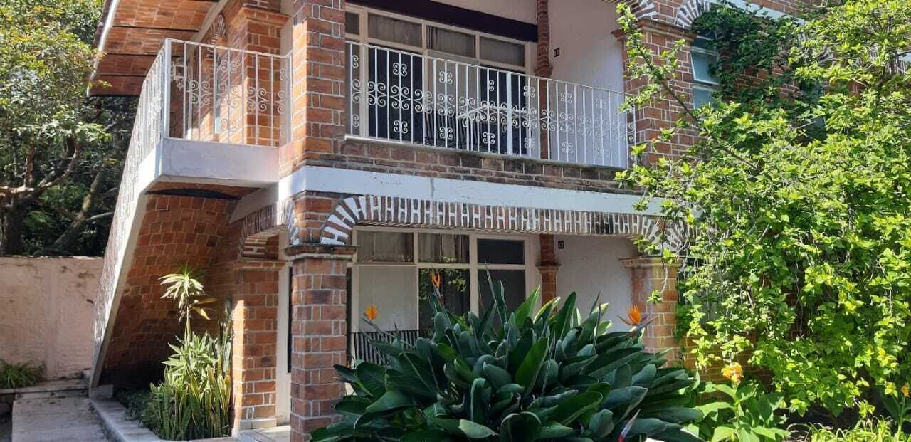 Un edificio de dos pisos con balcón, fachada de ladrillo y follaje circundante.