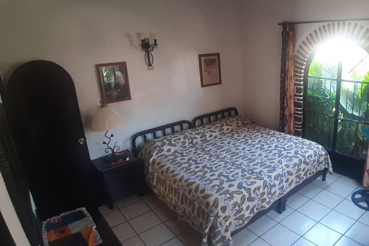 A bedroom with a bed, a nightstand, a wall-mounted lamp, a window with curtains, and a television.