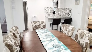 Un'area pranzo con un tavolo lungo, sedie a motivi floreali, una cucina sullo sfondo e un cartello che indica un ristorante.