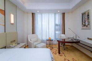 Habitación de hotel con cama, escritorio, silla, mesita con lámpara y un cuadro en la pared.