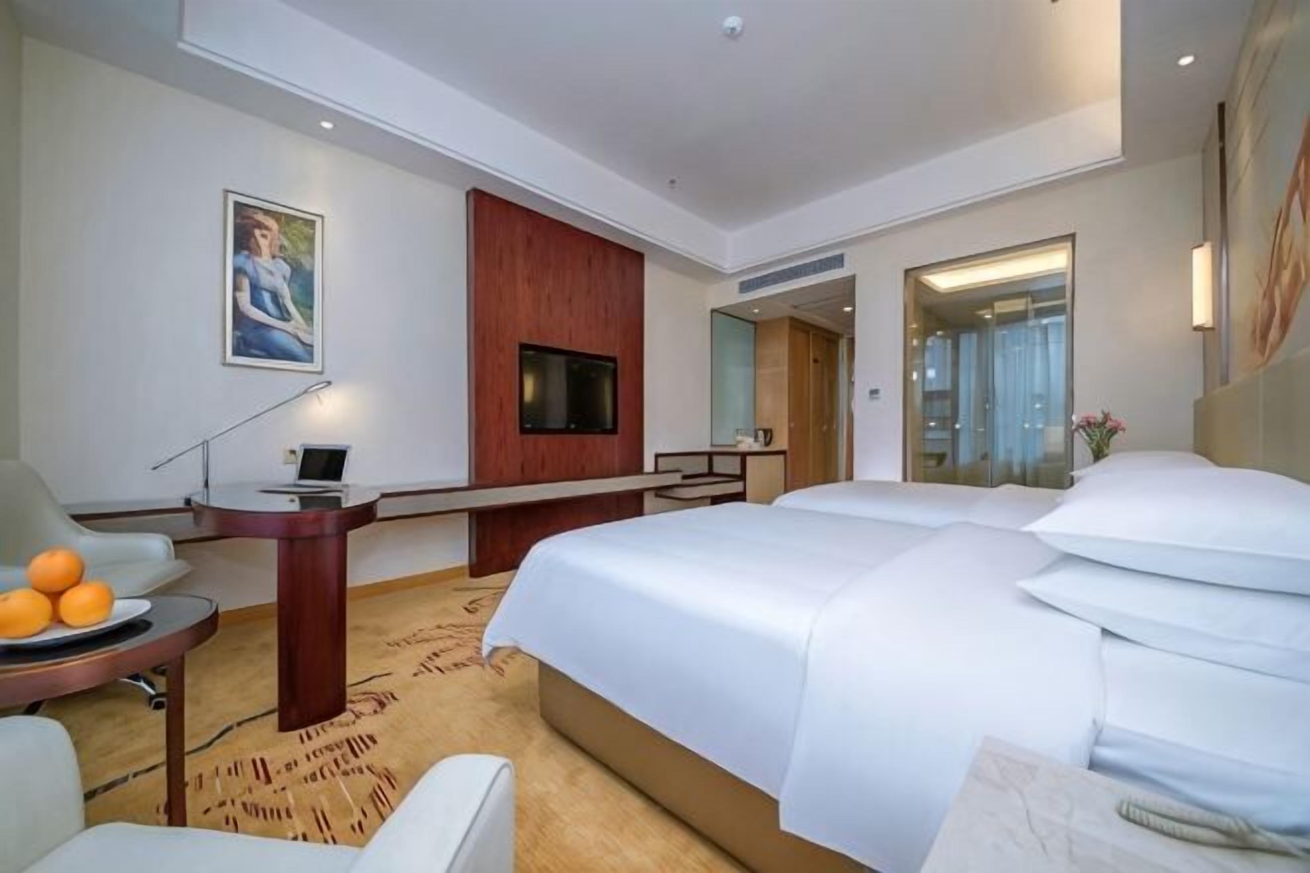 Hotellihuone, jossa on suuri sänky, työpöytä kannettavalla tietokoneella, tuoli, televisio ja taulu seinällä.