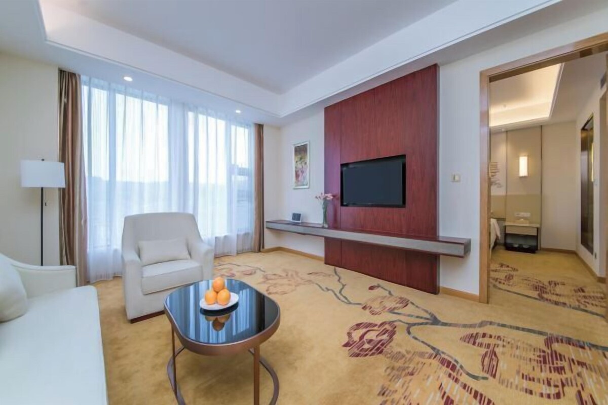 Habitación de hotel con cama, sofá, una mesa de centro con fruta, televisor montado en la pared y piso alfombrado.