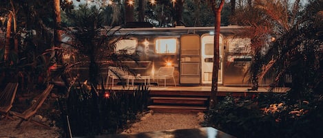 A stationary RV with a small porch, surrounded by dense foliage and string lights.