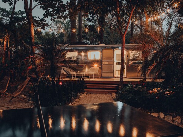 A stationary RV with a small porch, surrounded by dense foliage and string lights.