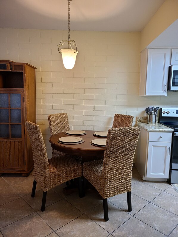 A dining area with a round table, four woven chairs, a hanging light fixture, a wooden cabinet, and a white microwave.