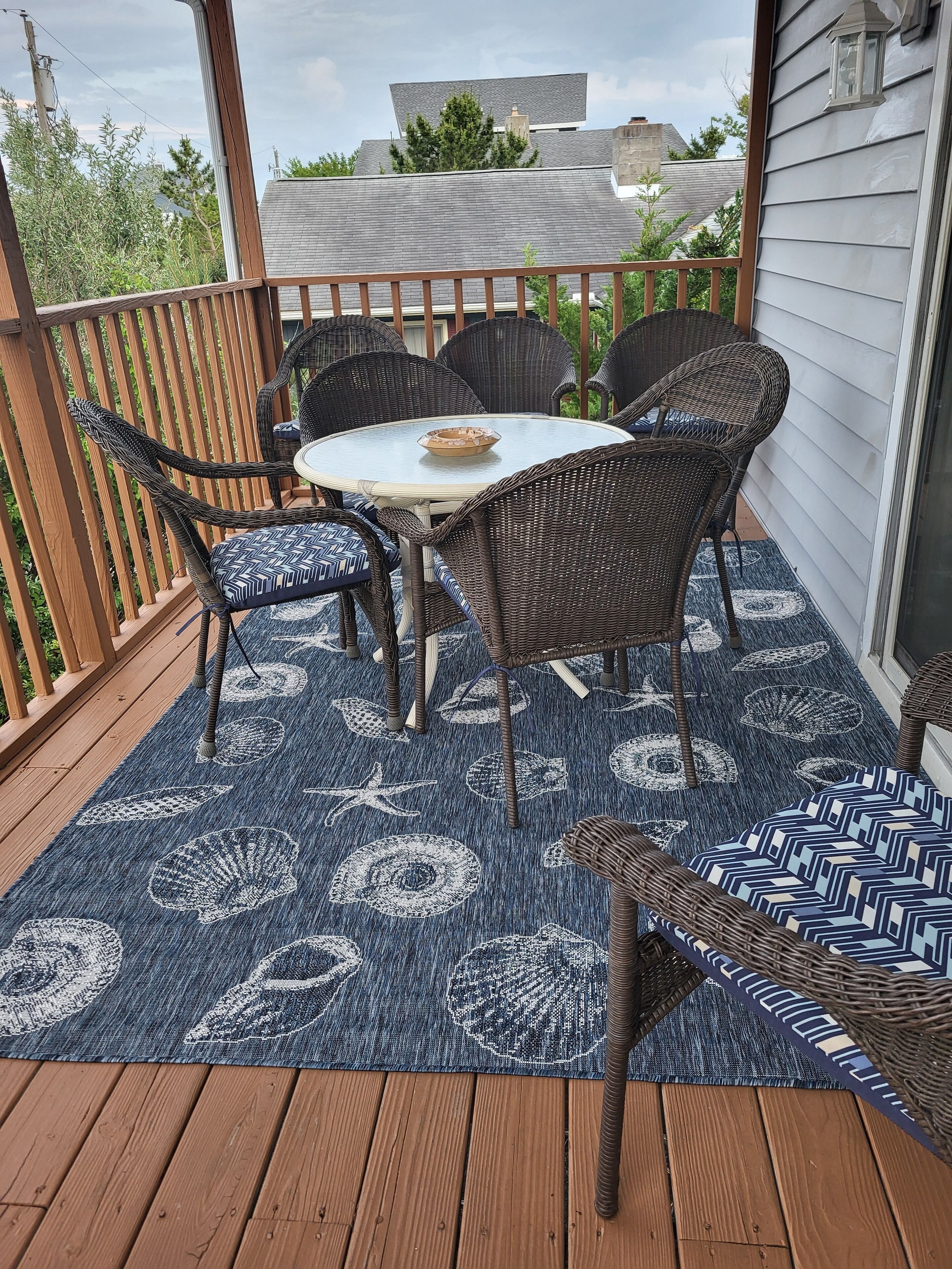 A covered outdoor patio with a round table and wicker chairs, a blue patterned rug, and a wooden deck.