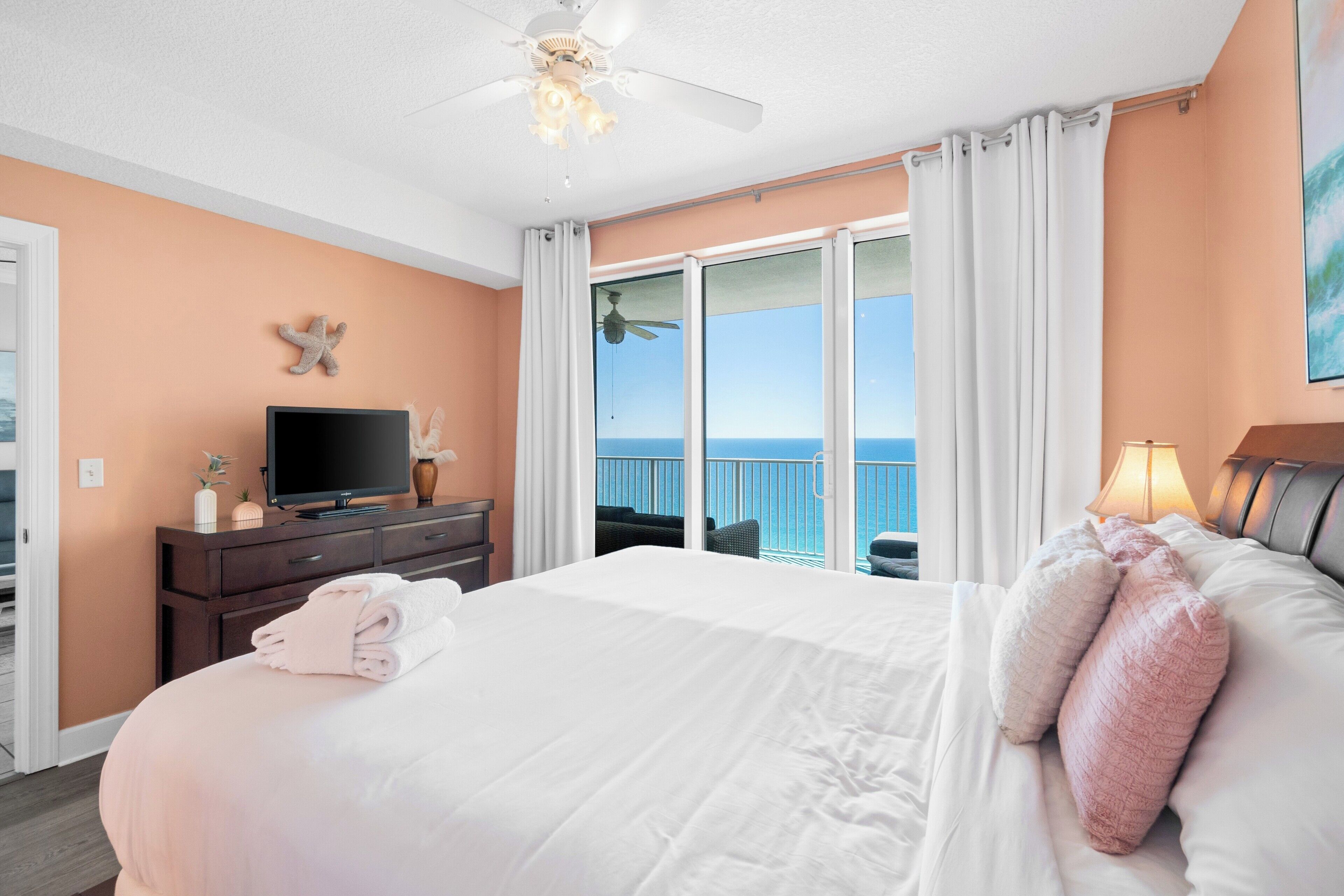 Habitación de hotel con cama, televisor, ventilador de techo, balcón con vista al océano y un cuadro en la pared.