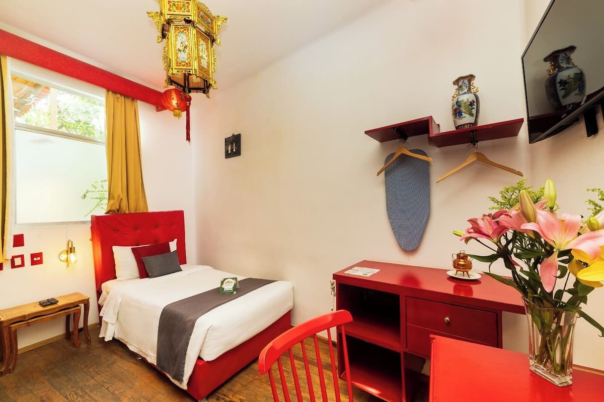 A hotel room with a red bed, a desk, a chair, a hanging lantern, and a vase with flowers.