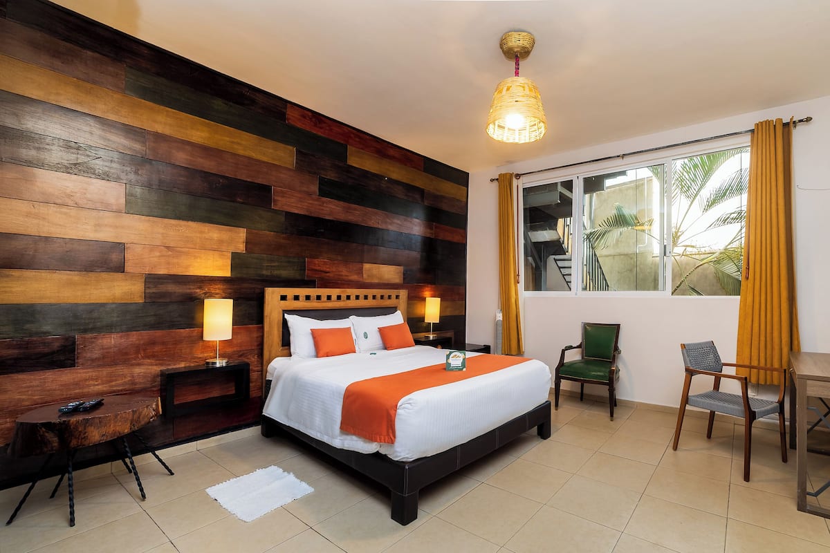 A hotel room with a wooden panel wall, a bed with white bedding and orange accents, two bedside tables with lamps, a chair, and a window with curtains.