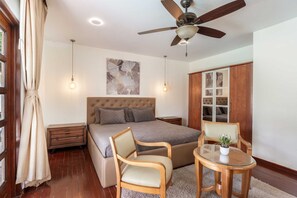 Ein Schlafzimmer mit einem Bett, einem Deckenventilator, einem Nachttisch, einem Sessel, einem kleinen Tisch mit einer Pflanze und einem Kleiderschrank.
