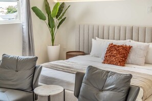 Un dormitorio con una cama, dos sillas grises, una mesita redonda y una planta en maceta.