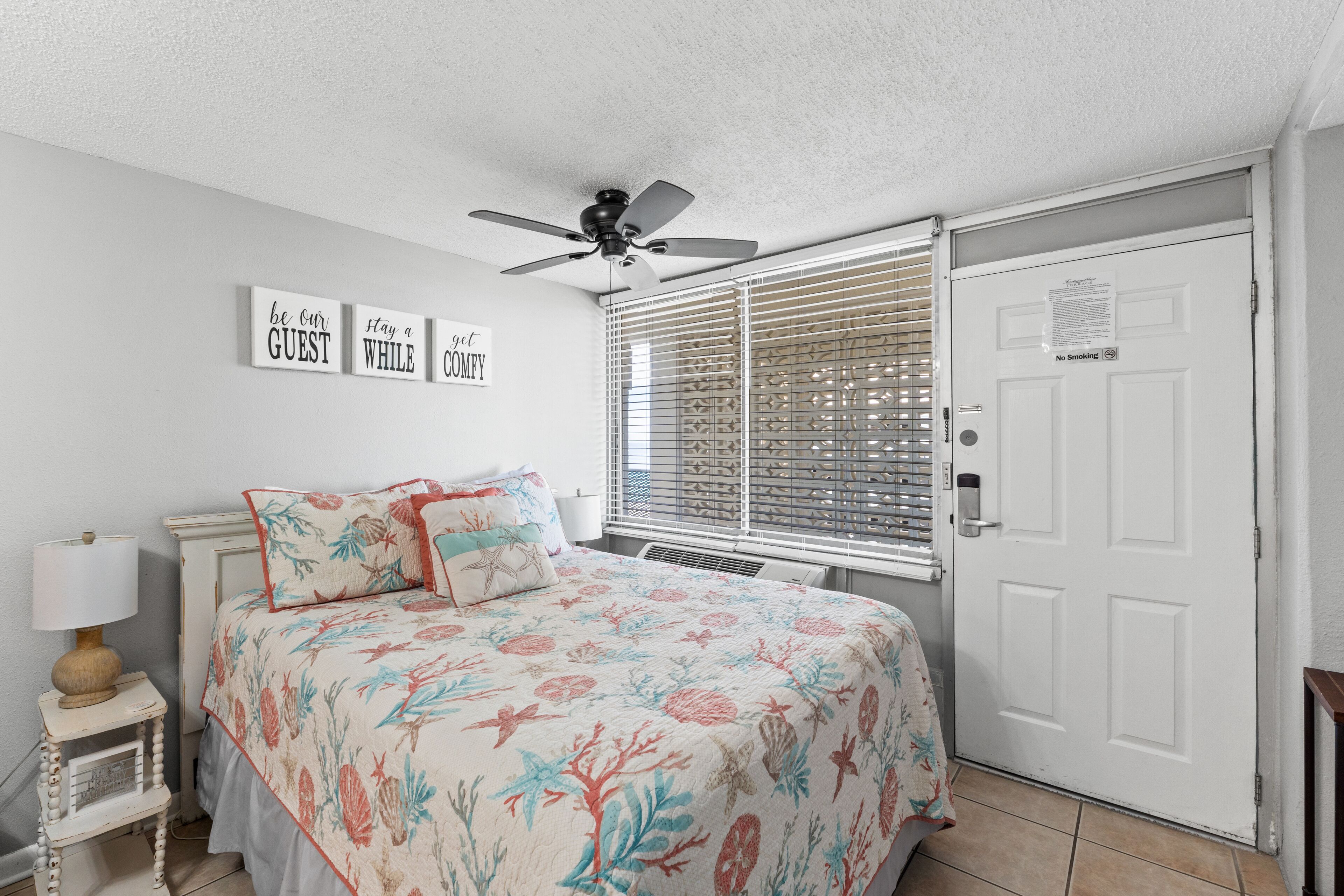 A bedroom with a bed, a ceiling fan, a window with blinds, a door with a 'No Smoking' sign, and wall decor with motivational phrases.