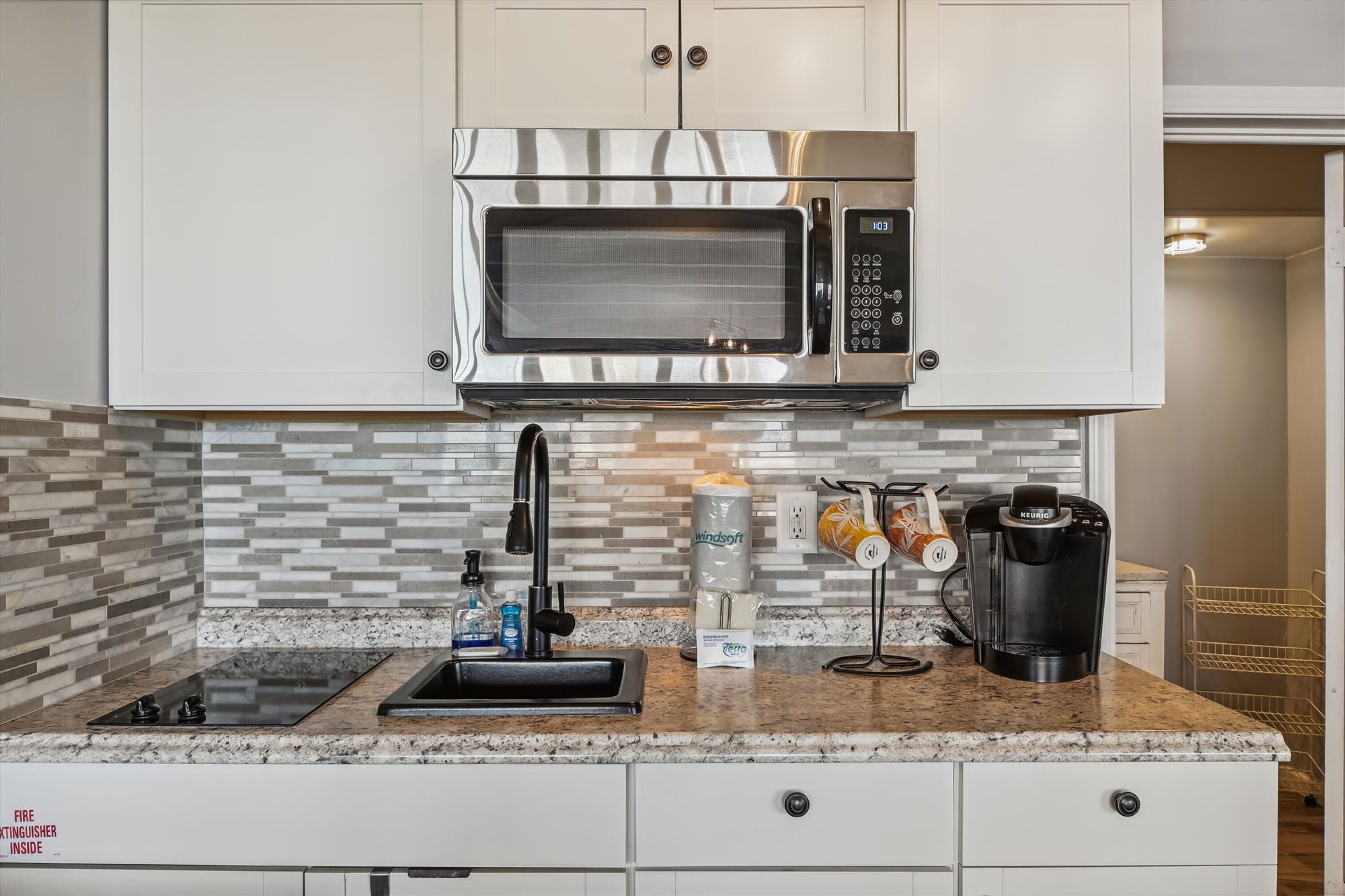 A kitchen with a stainless steel microwave, a sink, a coffee maker, and a tiled backsplash.