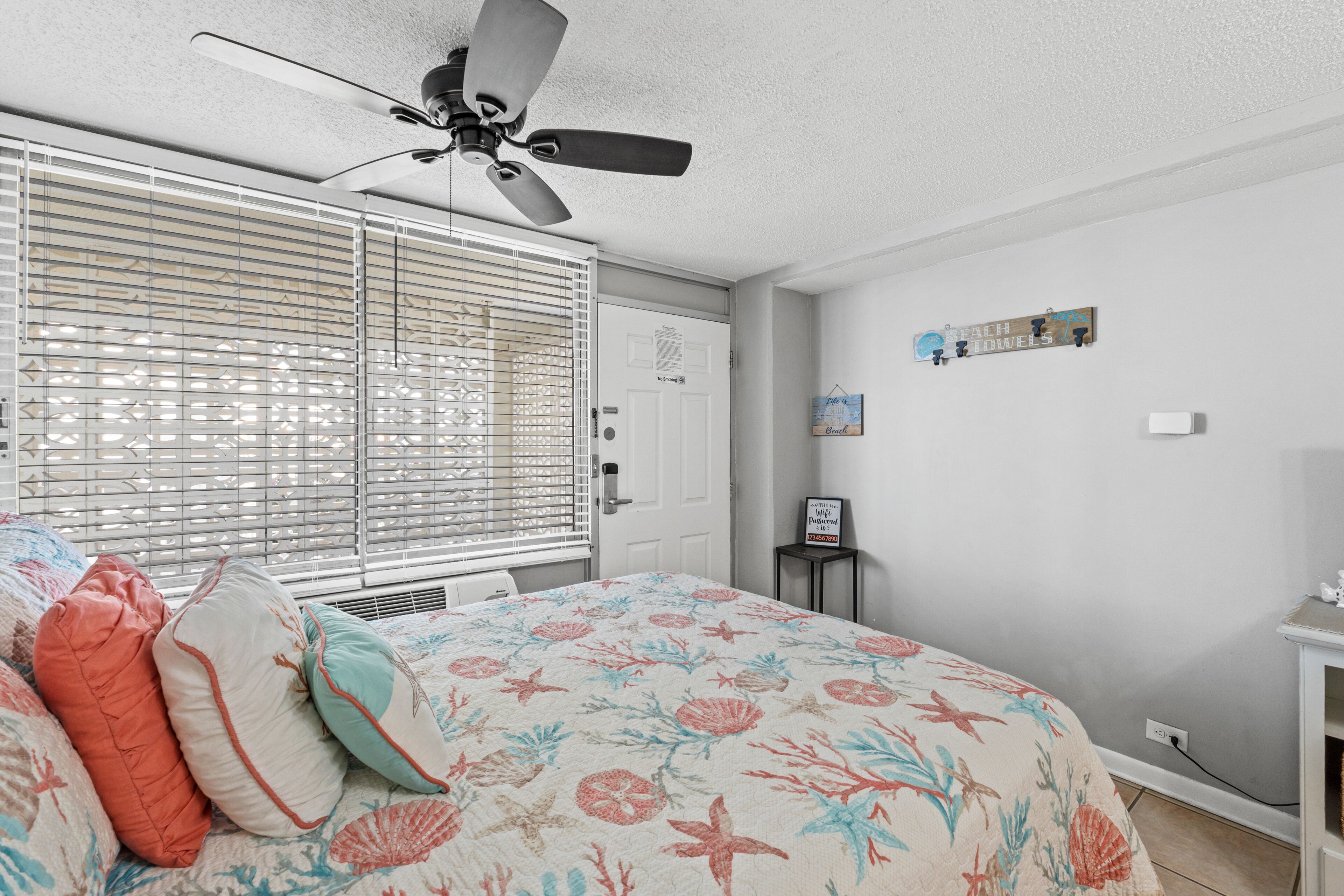 Ein Schlafzimmer mit einem Bett, einem Deckenventilator, einem Fenster mit Jalousien, einer Tür und einem kleinen Tisch mit einem Schild.