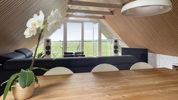 Un interior moderno con una mesa de comedor de madera, un sofá negro y sillas blancas. Se ve una ventana grande que ofrece vistas a un campo verde.