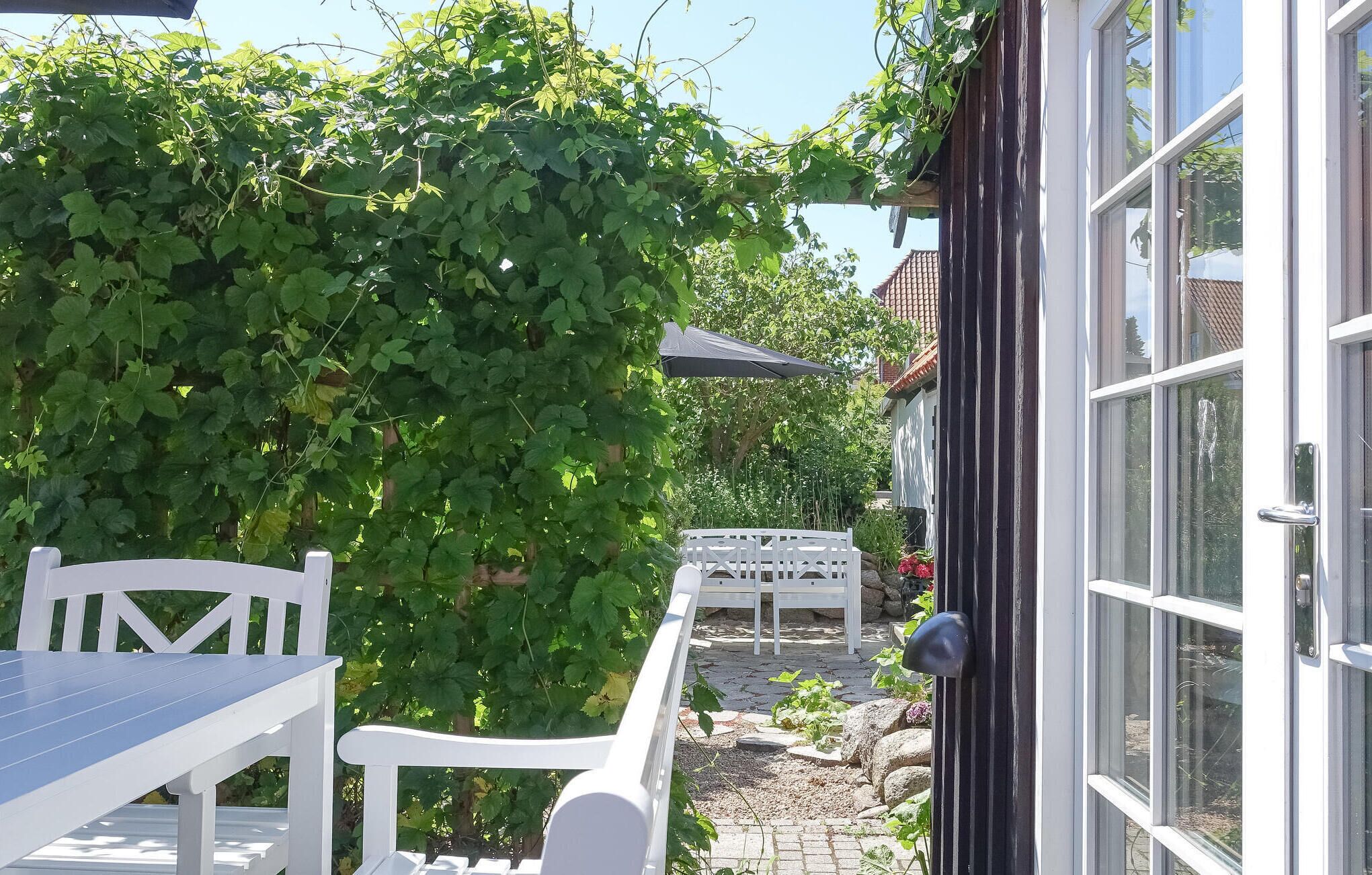 Eine Terrasse mit weißem Tisch und Stühlen, einer weißen Pergola und einer Glastür, die zu einem Garten mit weißem Zaun und viel Grün führt.