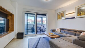 Una sala de estar moderna con un sofá, una mesa de centro y un televisor. Hay una puerta corrediza de vidrio que da a un espacio exterior con una mesa y sillas.