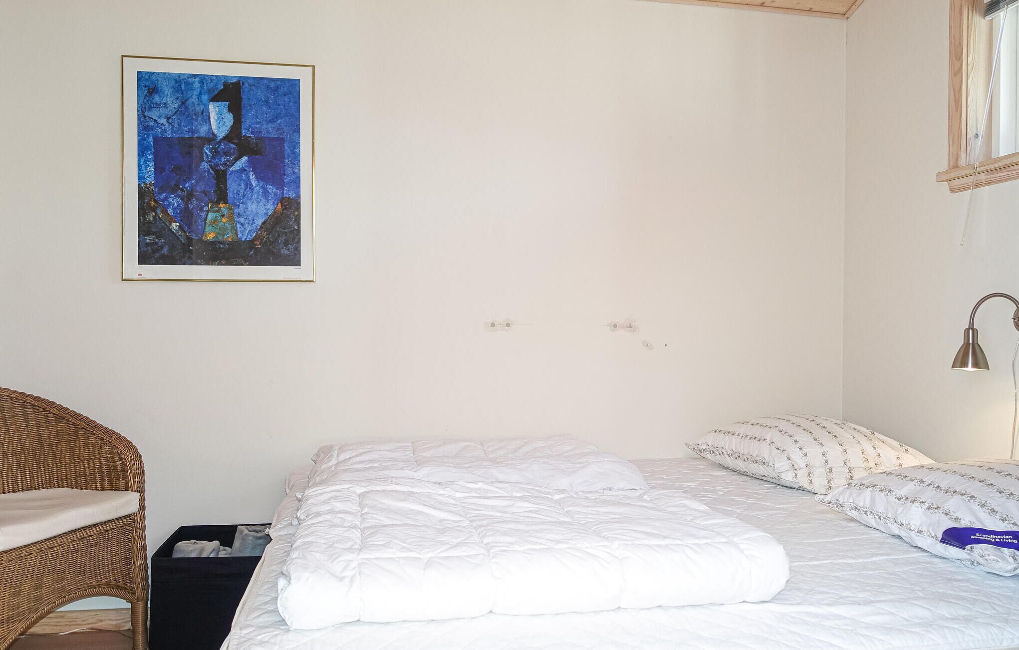 A bedroom with a bed, a wicker chair, a framed abstract painting, and a bedside lamp.