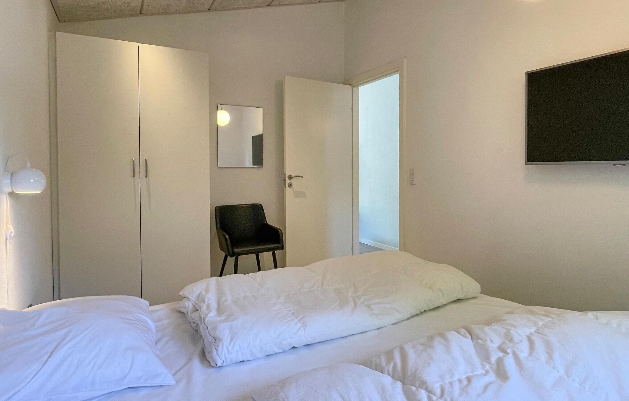 A bedroom with a bed, a chair, a wardrobe, and a TV on the wall.