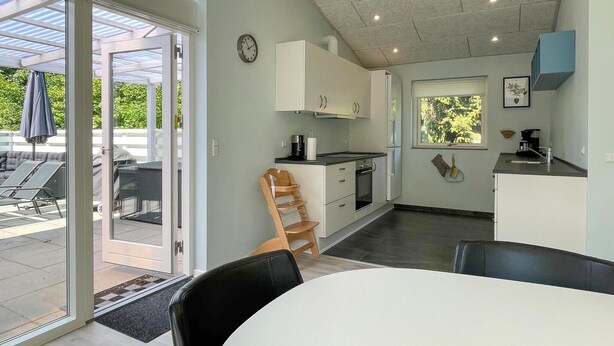 Una cucina moderna con mobili bianchi, forno integrato e una finestra. C'è un'area pranzo con tavolo e sedie, e un patio con barbecue e posti a sedere all'aperto.