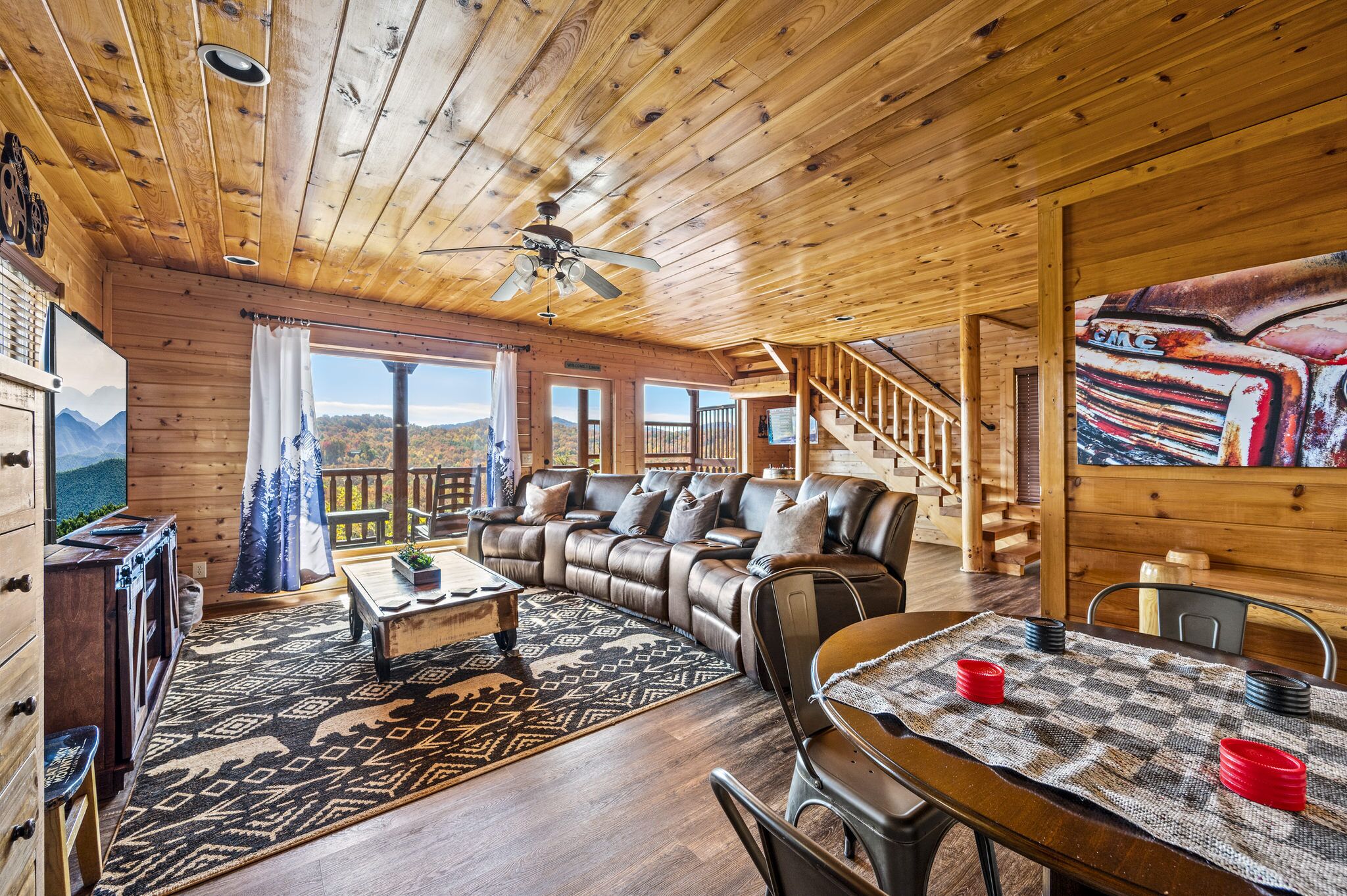 A wooden cabin interior with a sofa, dining table, and a view of mountains through large windows.
