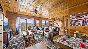 A wooden cabin interior with a sofa, dining table, and a view of mountains through large windows.