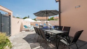En udendørs terrasse med et bord og stole, en pool med liggestole og parasoller.