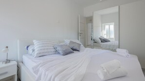 En pænt redt seng med hvidt sengetøj og grå puder, en sengetlampe og et sammenrullet håndklæde på gulvet.