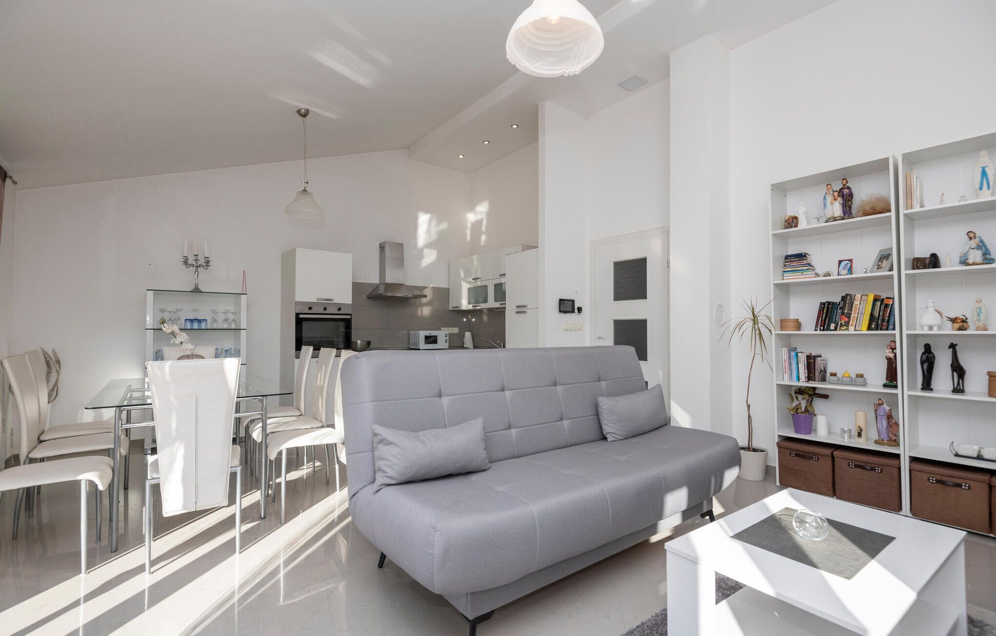 A modern living room with a grey sofa, a dining area with white chairs, a kitchen with white cabinets, and a bookshelf filled with books and decorative items.