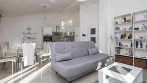 En moderne stue med en grå sofa, et spiseområde med hvide stole, et køkken med hvide skabe og en reol fyldt med bøger og pynteting.